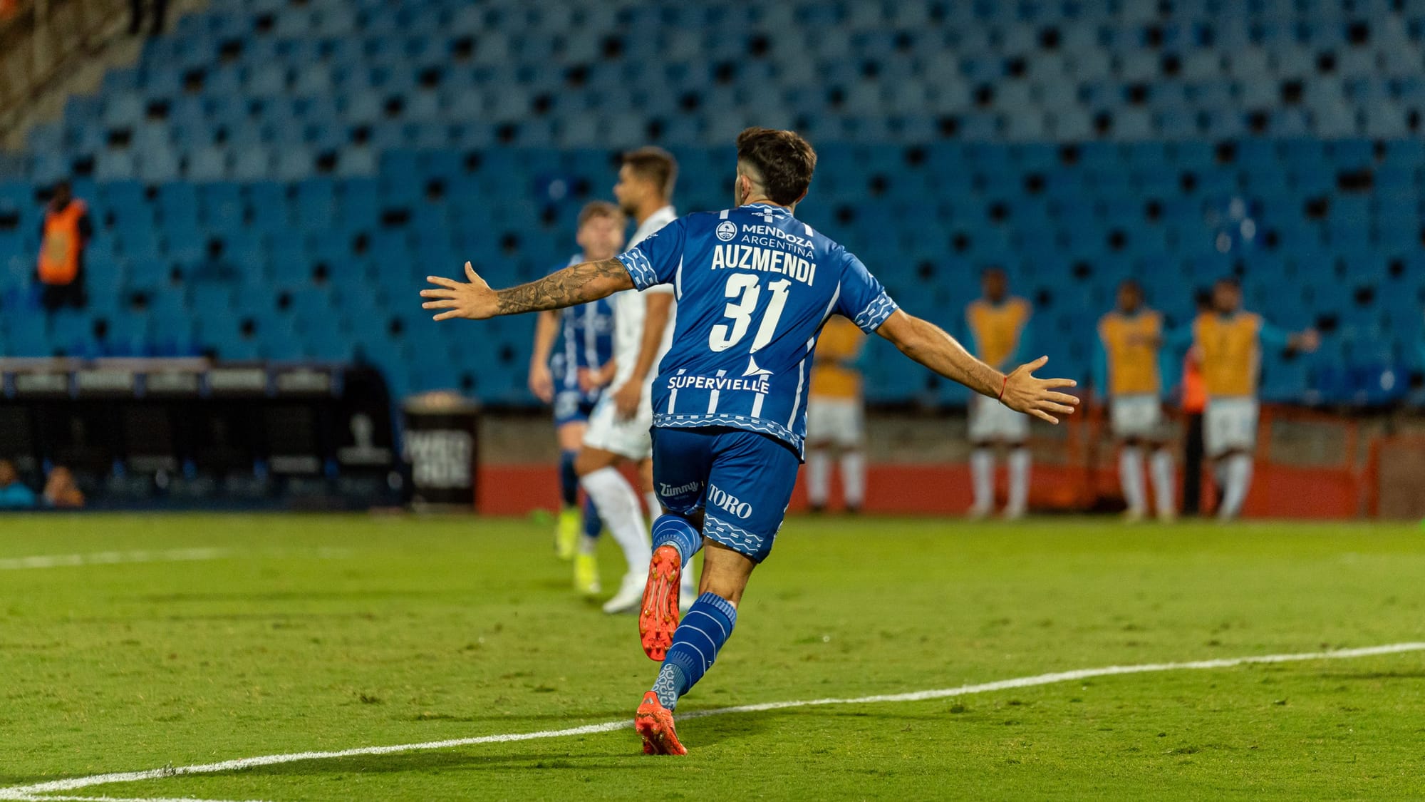 Jogador de futebol de uniforme azul comemorando gol de braços abertos no estádio